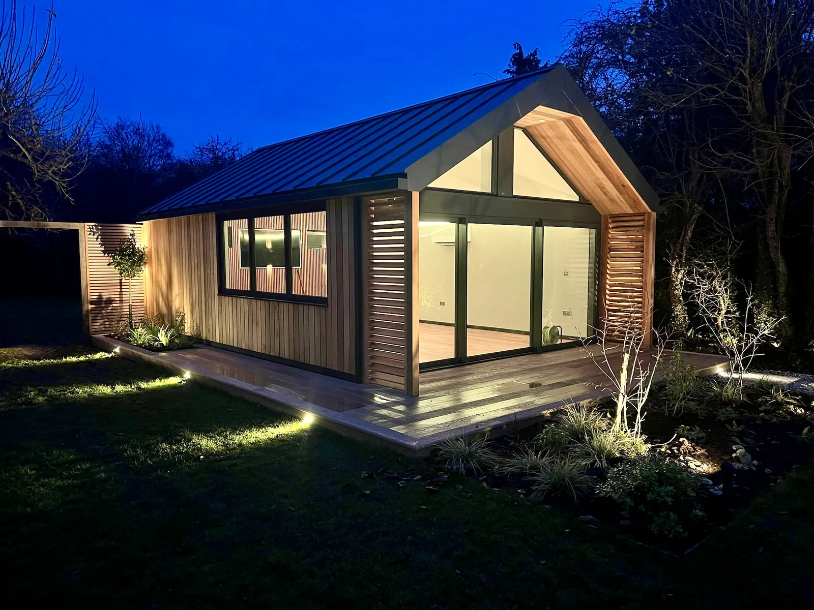 Timber-clad insulated garden room lit at dusk with landscaped decking, built by Helps & Co